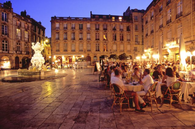 An evening scene with an illuminated old town square. There are people sitting at tables and a multi-story old building in the background.