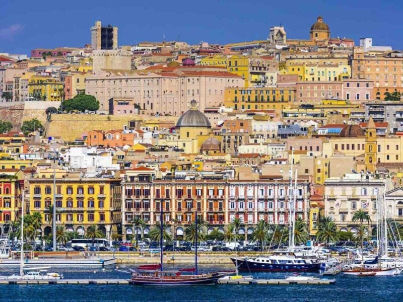 An old town sits on the water's edge, the town fills the image, rising up a hillside. There are boats in the water.
