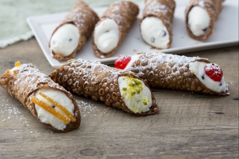 Three filled canoli lie on a table with four plated behind.