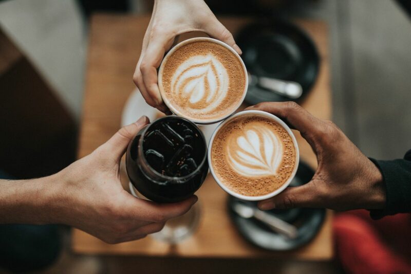 Photo taken from above three hands that are holding a cup of coffee each in the centre of the photo. The background is blurred.