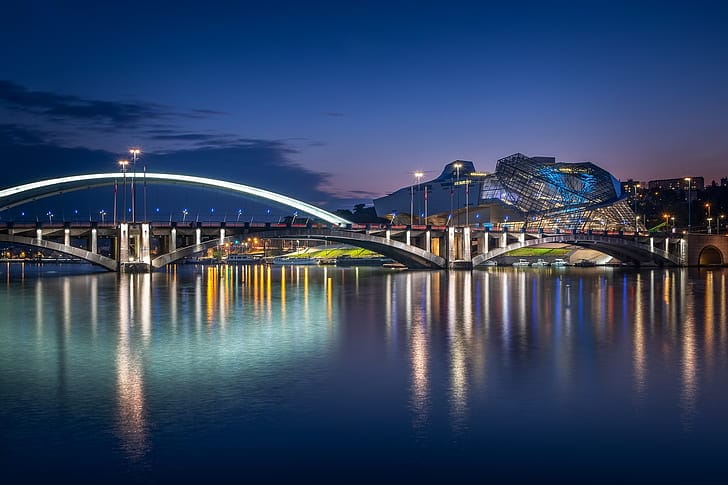 A modern bridge spans a wide section at the confluence of a river. The sky above is dark, perhaps pre-morning or early evening.
