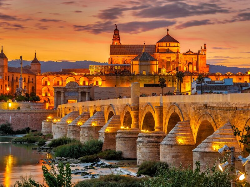 A golden sunset scene of Cordoba from across the rive with an old bridge in the foreground.