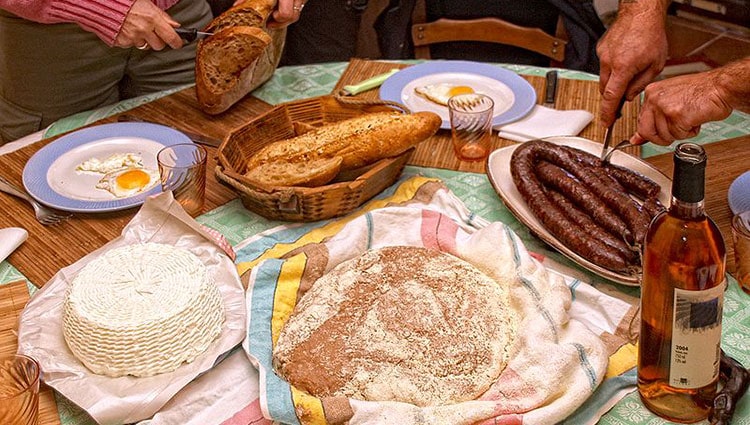 A table set with traditional Corsican food and a bottle of white wine, on a table cloth. Two people are partially visible; one is cutting a loaf of bread, the other cutting an Italian sausage.