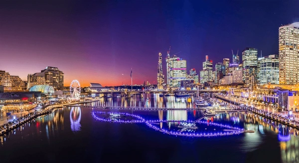 An evening scene of a harbour taken from across the water. The buildings are illuminated.