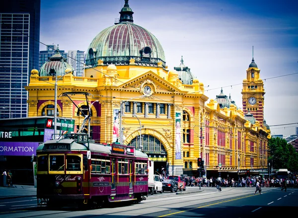 A large heritage train station is the background to a traditional Melbourne tram.