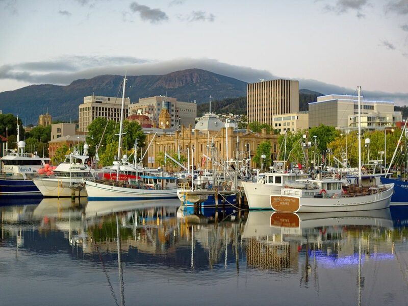 A city scene set behind boats floating on calm water. A large mountain in the background with a cloudy sky.
