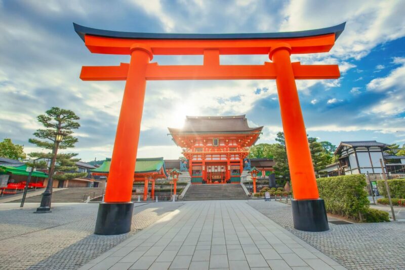 A traditional Japanese arch stands tall in the foreground. A straight path leads through the arch with steps leading up to a traditional Japanese building. There are various other buildings and trees surrounding the square.