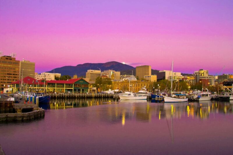 A city scene sits on and is reflected in a still body of water. There are boats on the water and a mountain in the background. The sky behind is purple and pink.