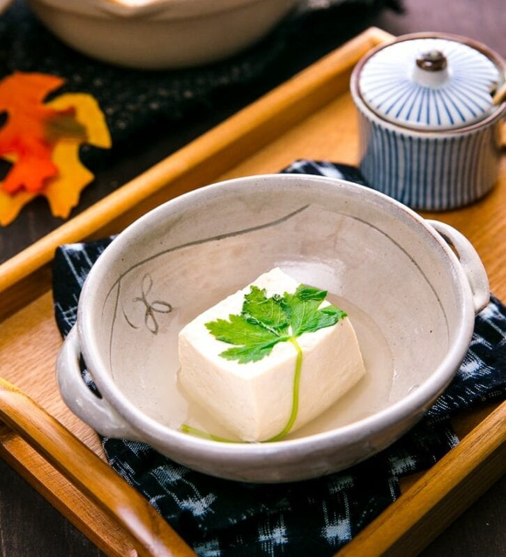 A single block of tofu topped with a green leaf sits in a bowl. The bowl sits on a napkin on a wooden tray with a small bowl with a lid behind.