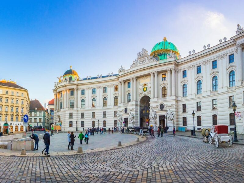 An old city square with cobble stones. Large impressive buildings surround the square; one with a green dome, another with a tall steeple.