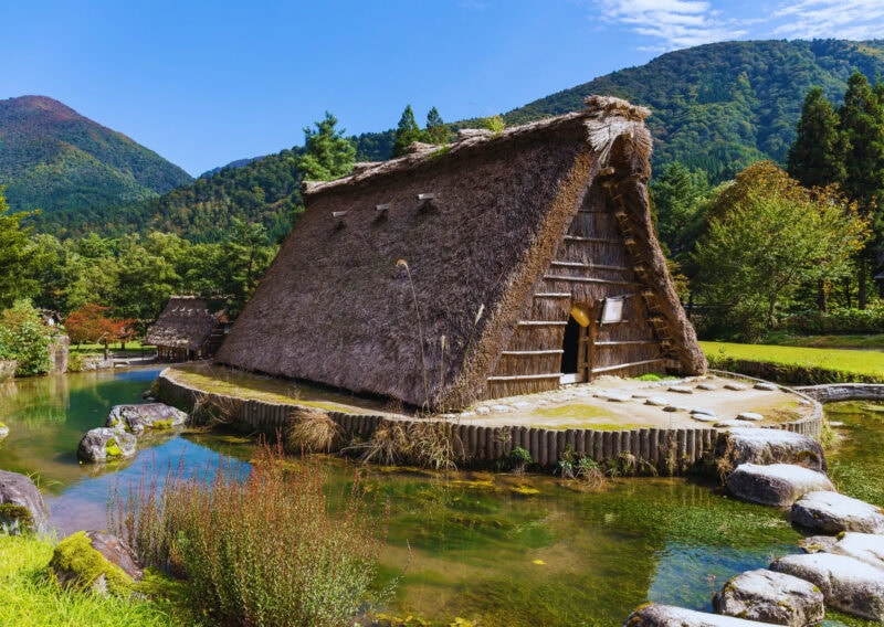 A traditional straw hut sits on the ground surrounded by a lake. There are stepping stones leading from the shore to the hut. There are hills in the background a blue cloudy sky behind.
