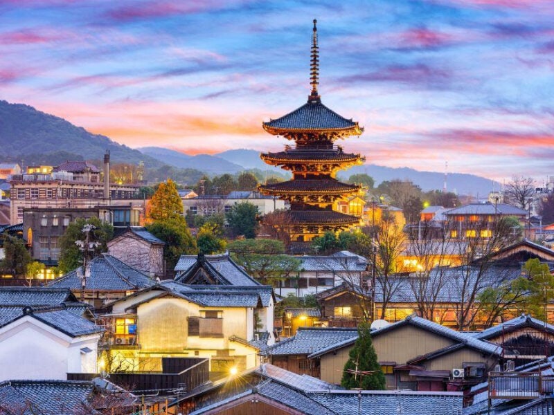 A traditional temple is the feature building in this city view of a Japanese old town with traditional Japanese buildings. Hills stretch across the background under a softly lit sky.