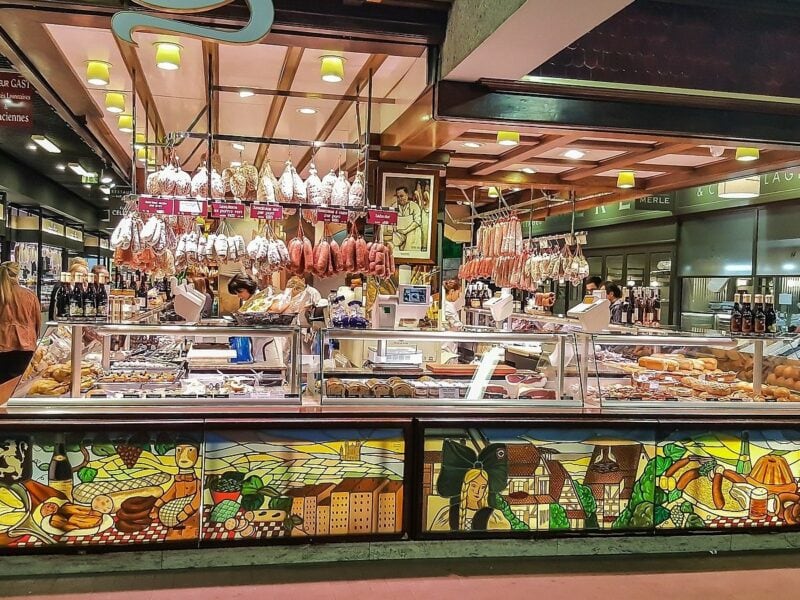 An undercover market scene with a meat shop featured in the centre. Other shops line the outside walls of the building.