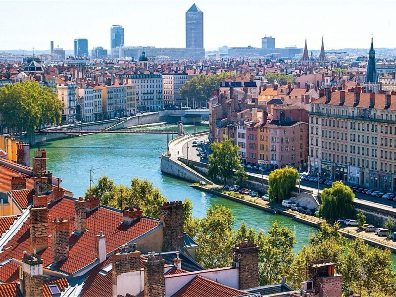 A view of the old town in a large city. There is a river winding through the centre of the image, with buildings and city scape trees along the banks. Tall buildings of the new city can be seen in the background.