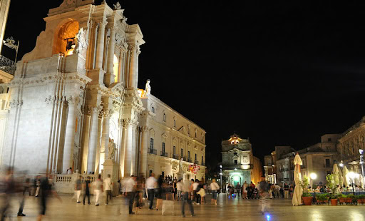 An Italian town square at night, surrounded by illuminated old town buildings. People wander the square.