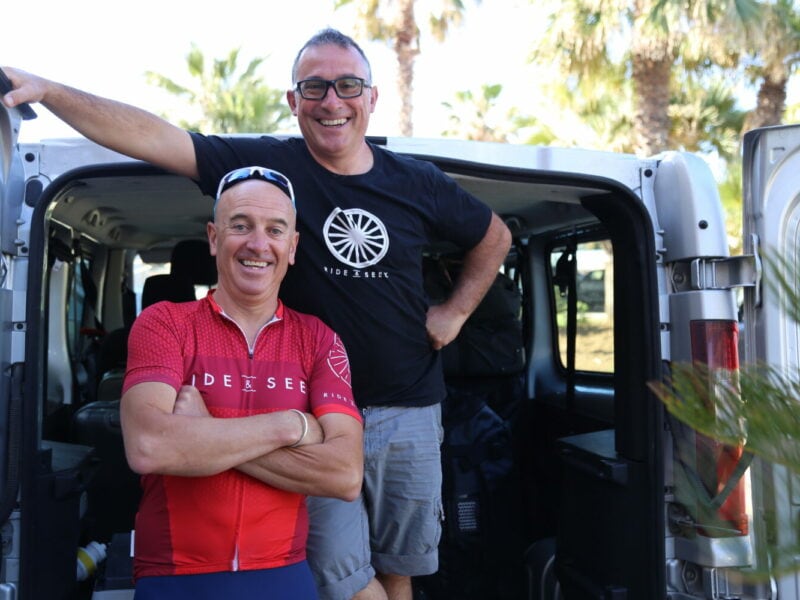 Two smiling guides wearing Ride and Seek uniforms pose naturally in front of the back of an open tour support van.