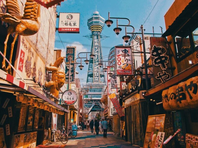 A narrow pedestrian street in a Japanese old town precinct, lined with traditional old shop fronts and lanterns. A tall modern tower stands tall in the background agains a light blue sky.