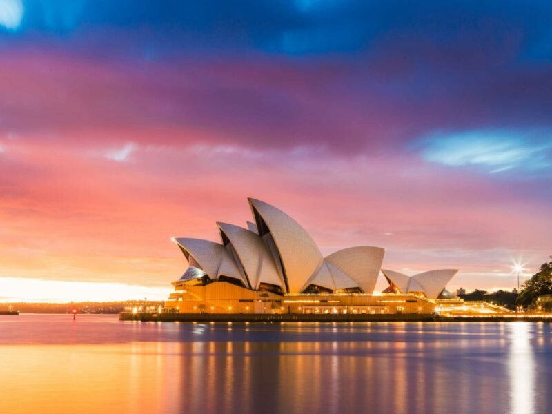 The Sydney Opera House is photographed from across the Harbour, with a colourful cloudy sky in the background.
