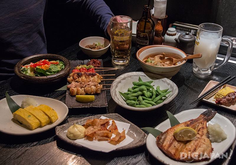 An array of dishes of dinner food are laid out in various styles of plates and bowls on a flat surface. A person's body is visible, holding a large glass of beer. There is also a beer bottle and some condiments on the table.