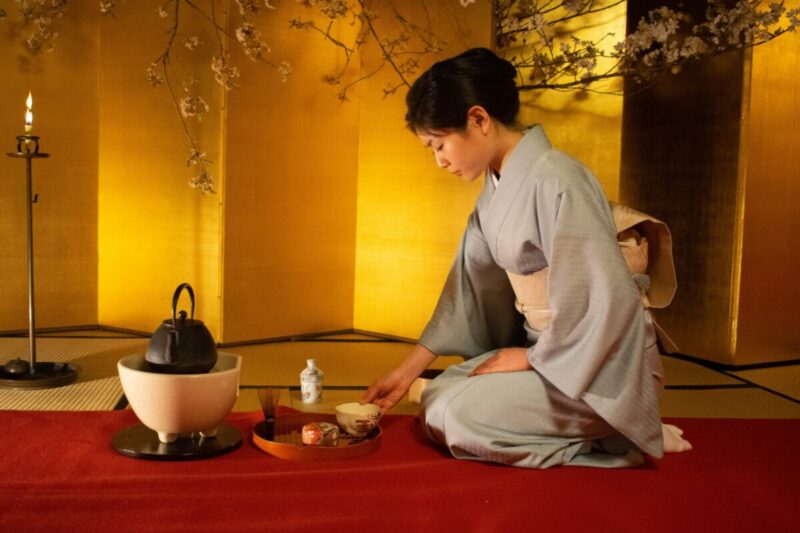A person in a yellow walled room wearing traditional Japanese clothing, kneels on a red carpet looking down. On the red carpet in front of her sits various items for the tea ceremony. There is a candle burning in a tall, thin, dark metal candle holder.