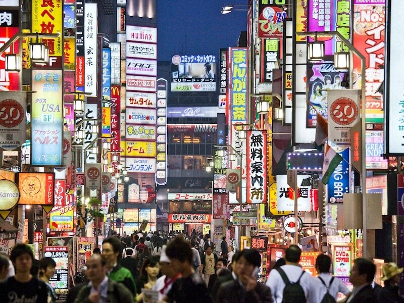 A city scene filled with tall buildings lit up with many coloured lights. Crowds of people walk the street at night time.