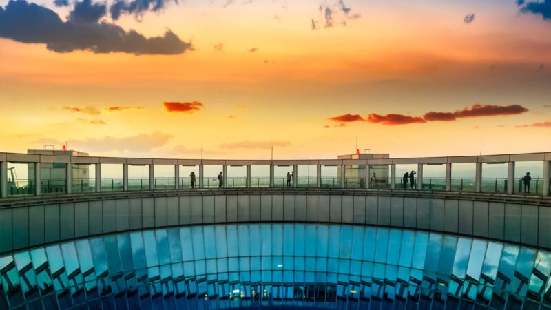 The top of a skyscraper, a round building with mirrors. There is an orange coloured sunset sky behind. People are standing in the building.