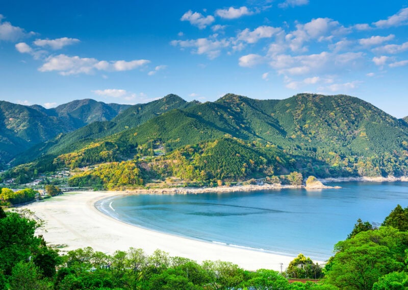 A bay with a clean white sandy beach. There are mountains in the background with a cloudy blue sky behind.