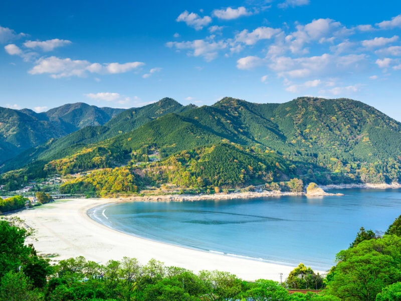 A bay with a clean white sandy beach. There are mountains in the background with a cloudy blue sky behind.
