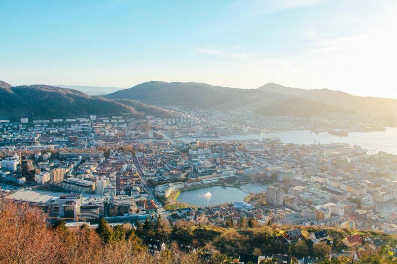 A view of a city from afar. There is a small lake and an inlet within the city area shown. A range of hills are in the background with a blue sky behind.