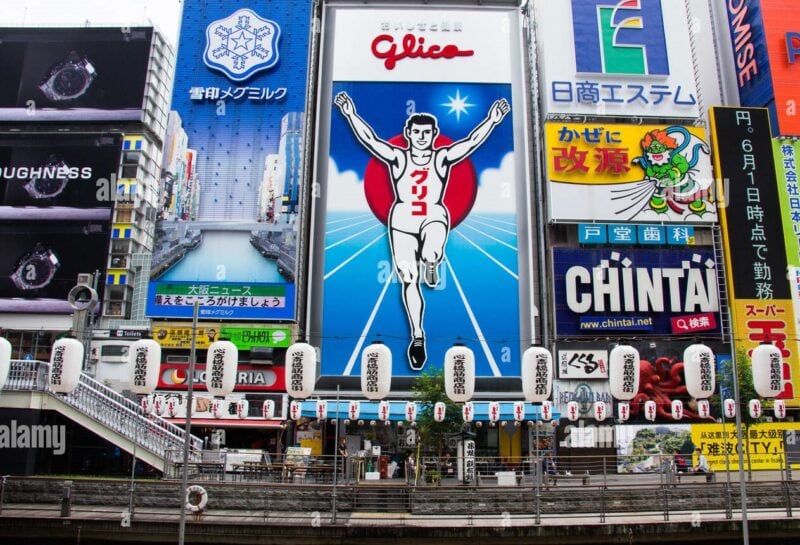 A large mural of a man in sports clothing runs with his hands stretched upwards. The billboard sits amongst a variety of other large, colourful billboards on buildings on a street lined with Japanese lanterns.