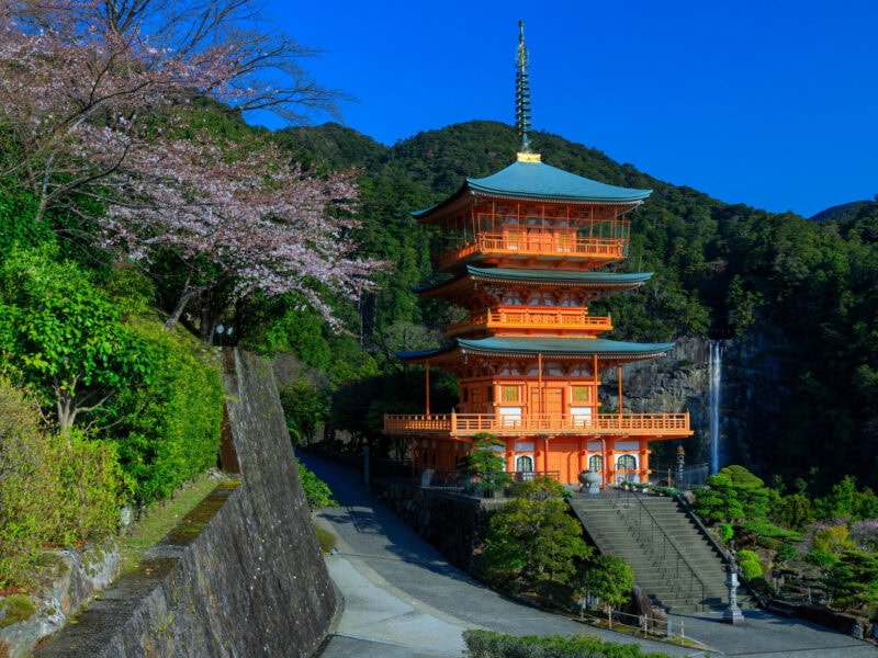 A multi-story traditional style Japanese temple sits part way up a steep hill. Stairs lead from a path running beside the temple up to the entrance. A waterfall on the right and a tall hill in the background with a clear blue sky behind.