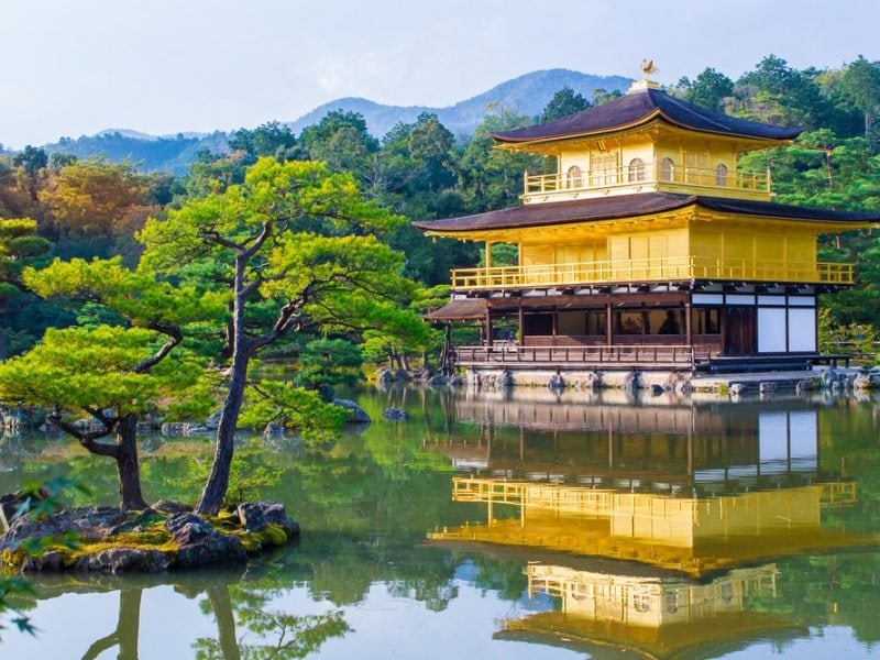 A traditional Japanese building of two stories sits on the edge of a lake that is surrounded by trees. The scene is reflected in the still water of the lake in the foreground.