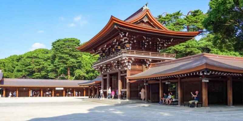 A traditional Japanese shrine with a traditional roof sits in a wide open square. There are visitors sitting and walking in the shade. A small family are walking towards the entrance.