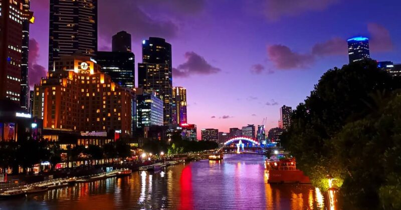 Modern city skyscrapers sit on the banks of a river. The night evening scene is illuminated, including a bridge at the far end of the river.