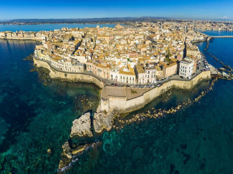 A view of an old town, perched on the clifftops, surrounded by blue water. The island stretches off into the background.