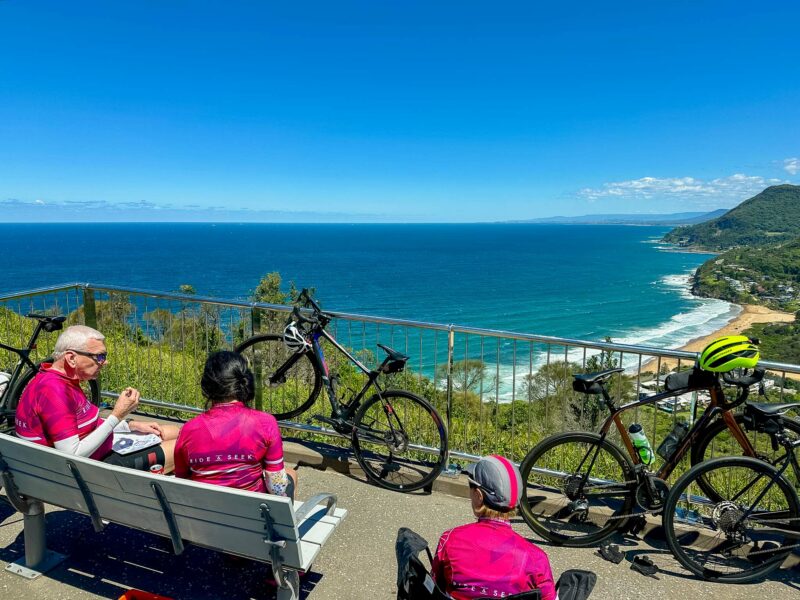 Cyclists in bright magenta Ride and Seek jerseys resting on a bench at Bald Hill Lookout, overlooking the sweeping Wollongong coastline and deep blue ocean.