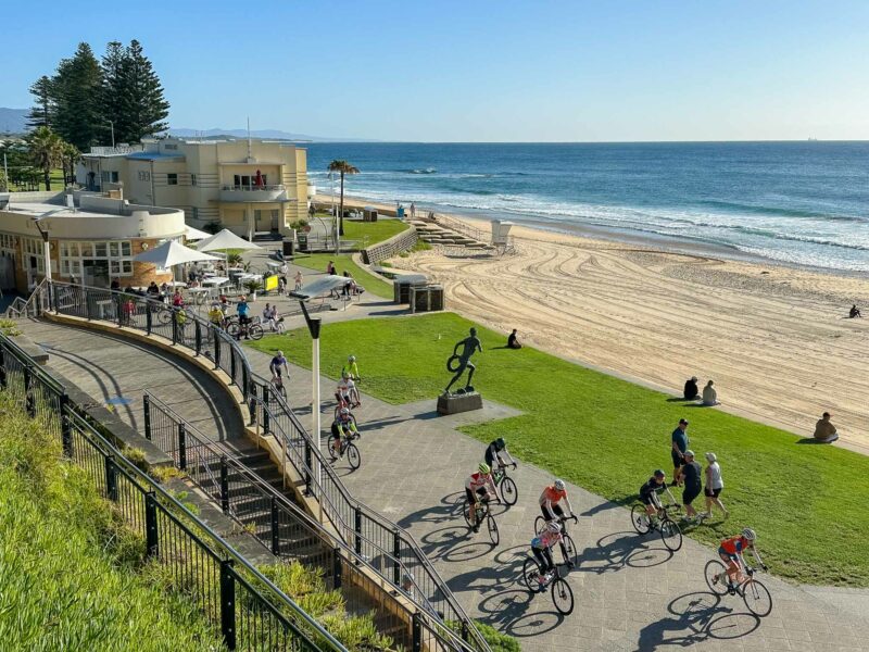 Group of riders cycling past a beachside pavilion and statue on a sunny day at a sandy foreshore.