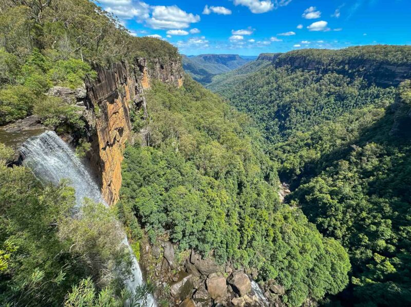 Waterfall cascading over a cliff into a deep forested valley with rugged sandstone walls under a bright blue sky.