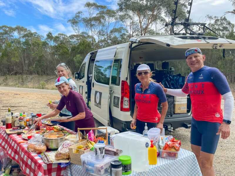 Group of cyclists gathered around a roadside picnic table covered in food, with support van parked nearby.