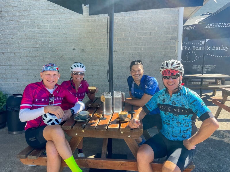 Cyclists enjoying coffee together at an outdoor café table at The Bean and Barley coffee shop, on a sunny day.