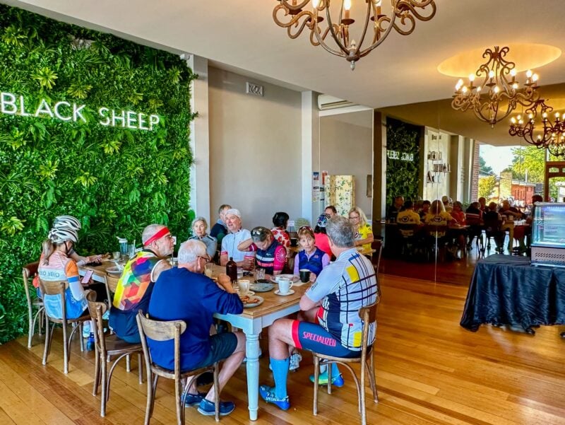 Large group of cyclists gathered for breakfast inside a café with chandeliers and a green wall reading ‘Black Sheep’.