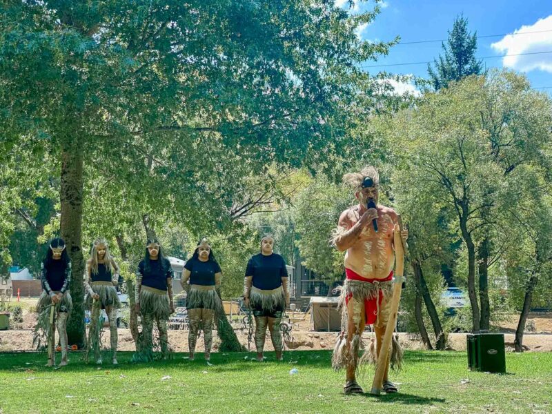 Indigenous Elder, Uncle Dozer in traditional body paint and attire addressing an audience, with a group of Indigenous women dancers in traditional body paint and attire standing behind him under the shade of the trees.