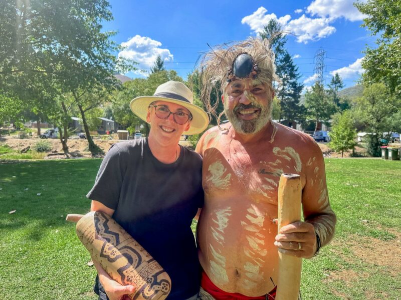 Smiling guide posing with Uncle Dozer, in traditional Indigenous body paint and headdress. Both are holding cultural instruments in a sunny park setting.