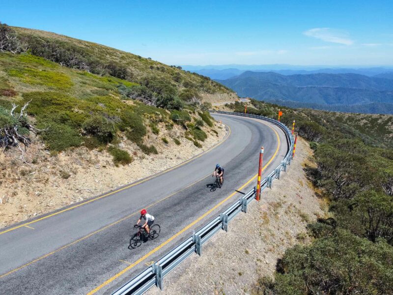 Cyclists climbing the Great Alpine Road on the ridge to Hotham Village with wide mountain views and a bright blue sky.