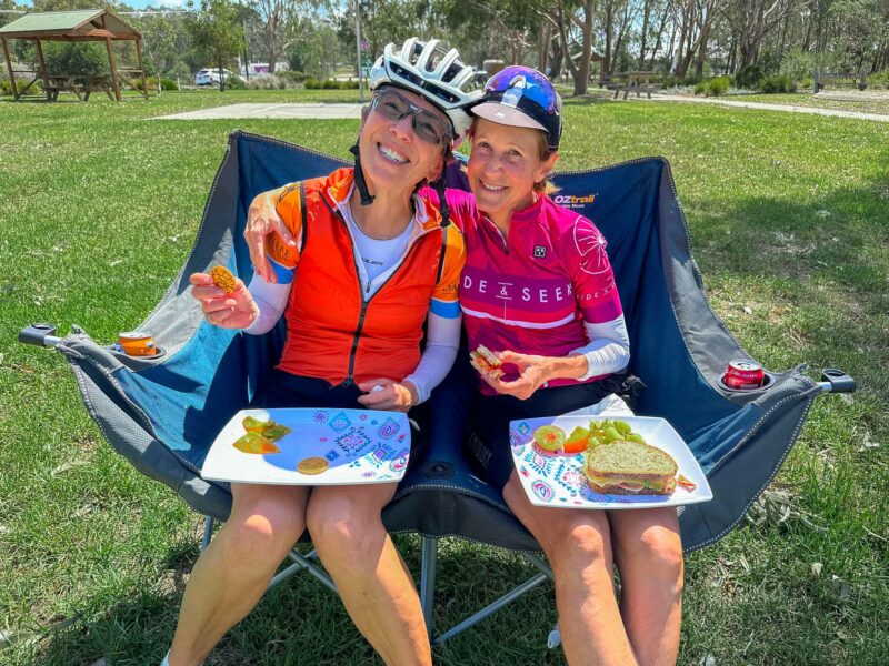 Two women in cycling jerseys sitting on camp chairs, smiling and enjoying sandwiches and snacks outdoors.