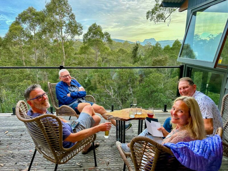 Group of friends relaxing with drinks on Cradle Mountain Lodge balcony overlooking a forest with distant mountain views at sunset.