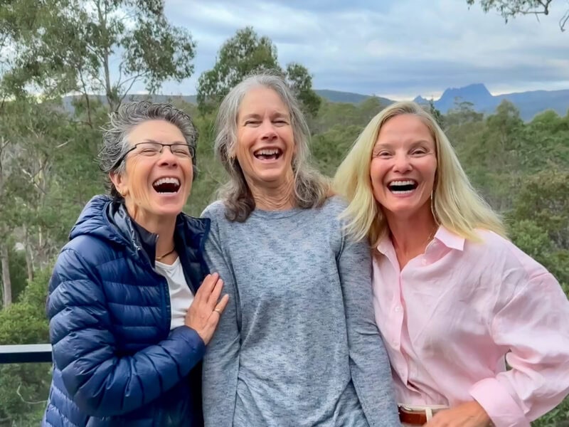 Three guests pose laughing on an outdoors balcony. Forest trees and Cradle Mountain in the background.