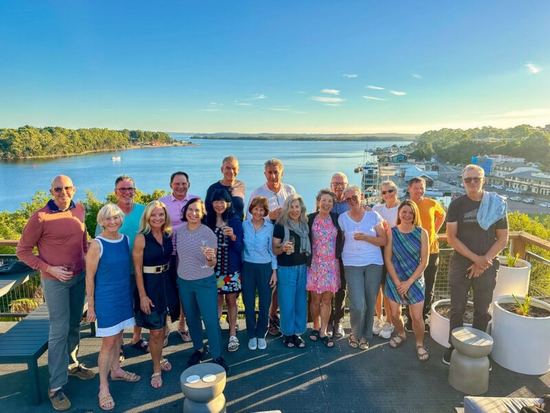 Smiling guests pose smiling in a group photo dressed for dinner, on the Stahan Village terrace overlooking the water and marina at sunset.