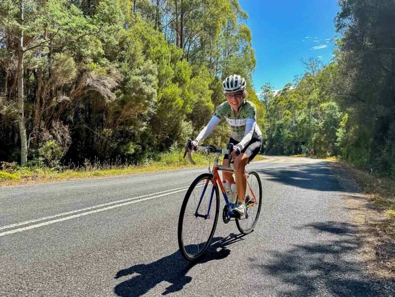Cyclist riding along a winding forest road lined with tall green trees and clear blue sky in the background.