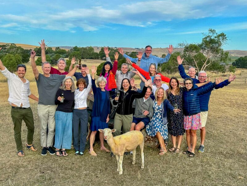 A large group of people standing in a field with a single sheep in the foreground. The group is smiling and waving at the camera, with rolling hills in the background.